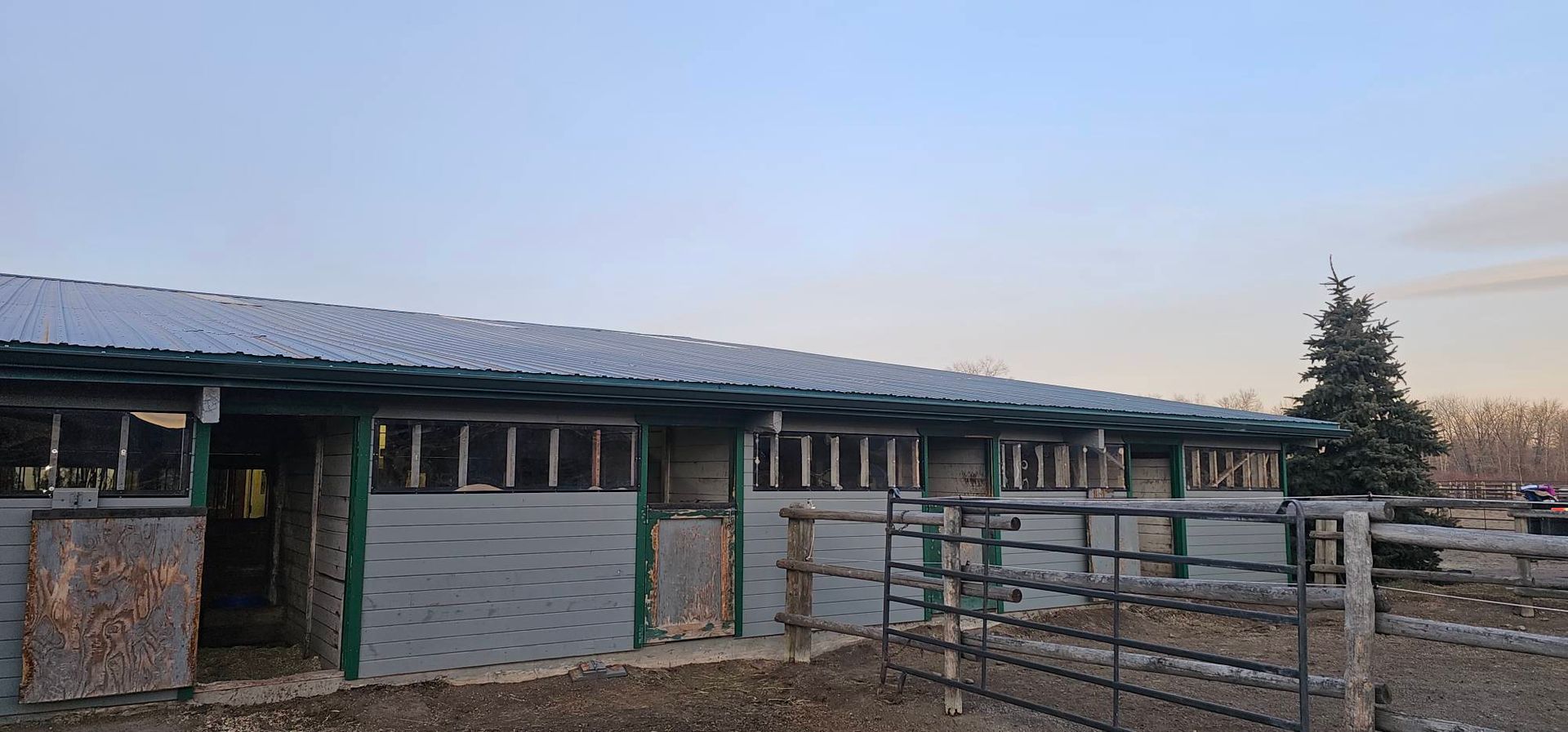 A long, single-story stable with a corrugated roof, gray walls, and a fenced outdoor paddock area at sunset.