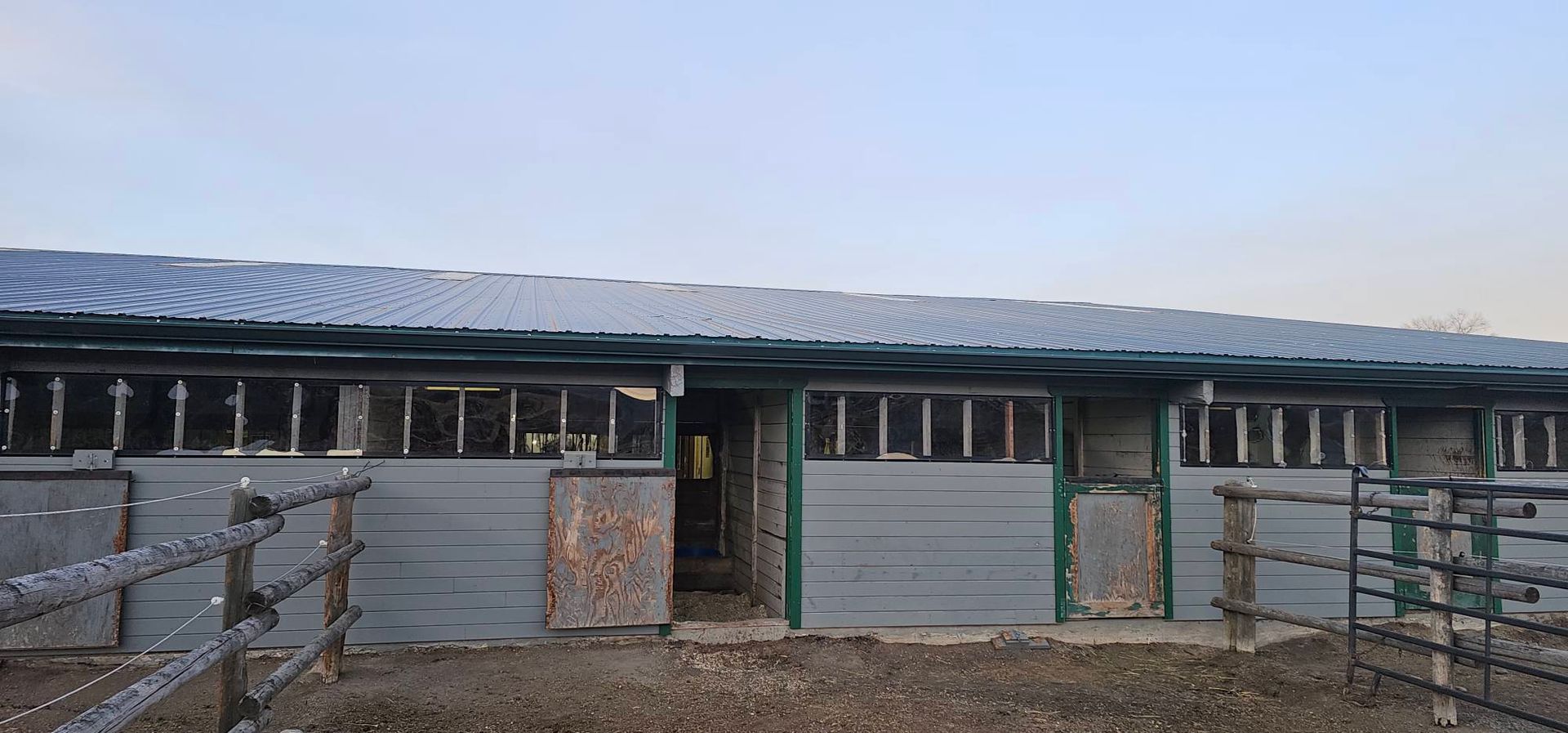 A gray, single-story stable building with multiple stall doors and windows, set behind a wooden fence in a yard.