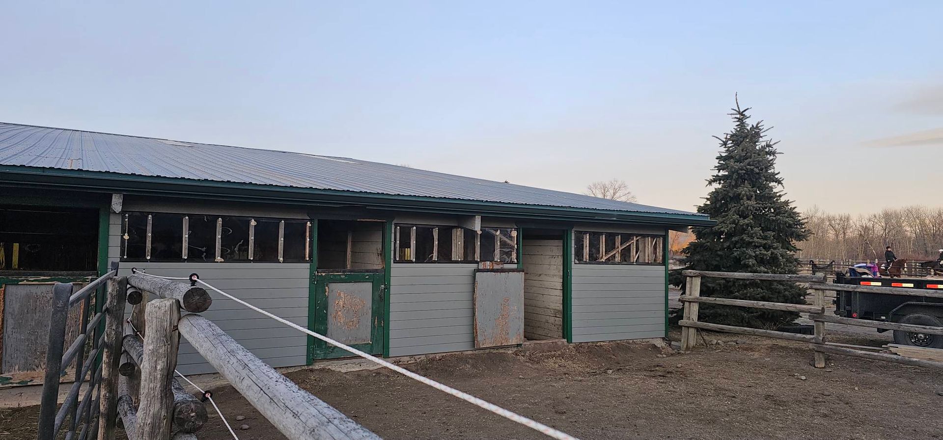 A long, grey-sided horse barn with dark trim, under a pale sky next to an evergreen tree in a dirt paddock.