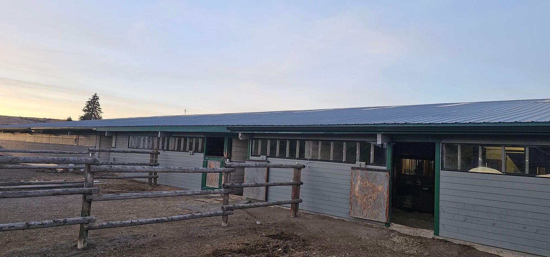 A long, single-story stable building with grey metal siding and a roof, featuring open stalls behind a wooden fence.