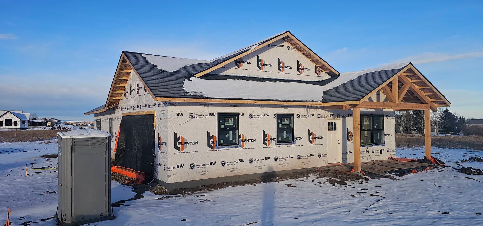 A partially built house with white protective siding, a wooden front porch, and black window frames in a snowy, open field.