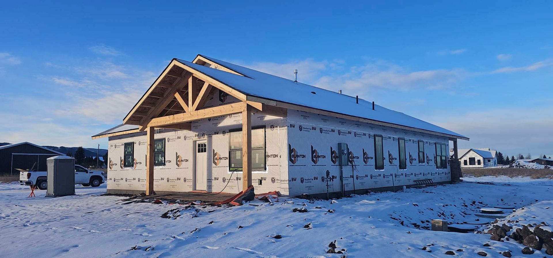 A newly framed house under construction with white house wrap, a wooden porch frame, and snow-covered ground.