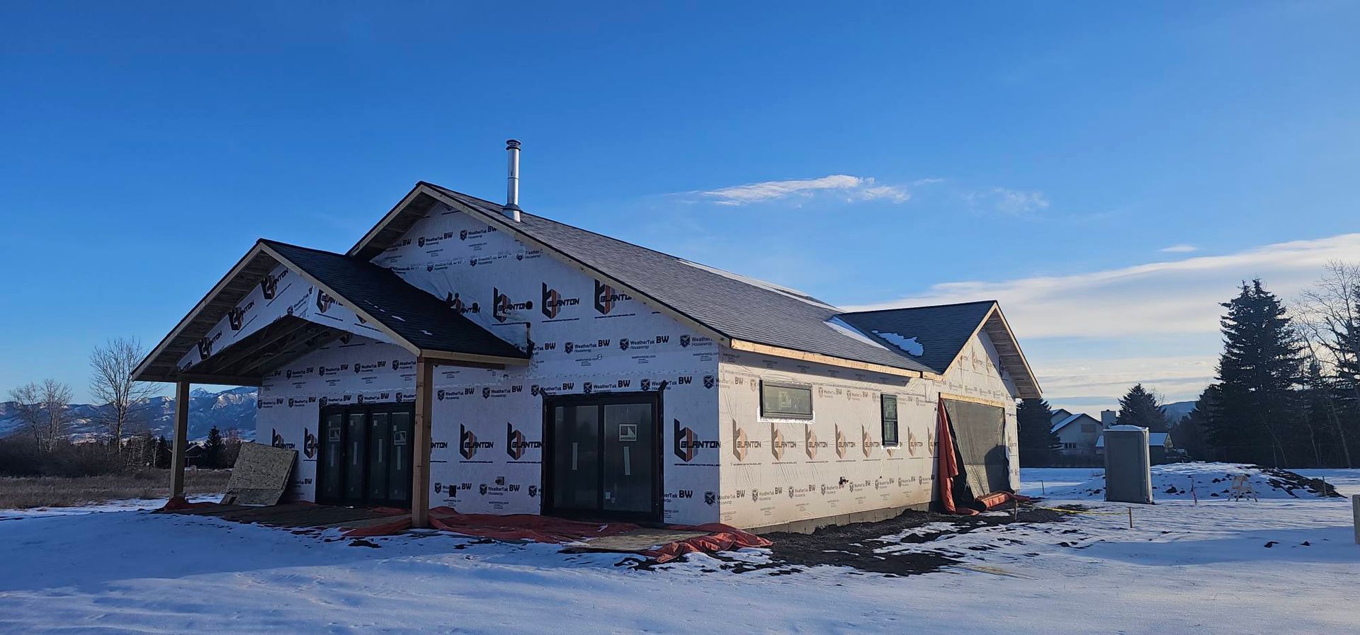 A house under construction with white exterior sheathing, a gabled roof, and glass doors, situated in a snowy, open landscape.