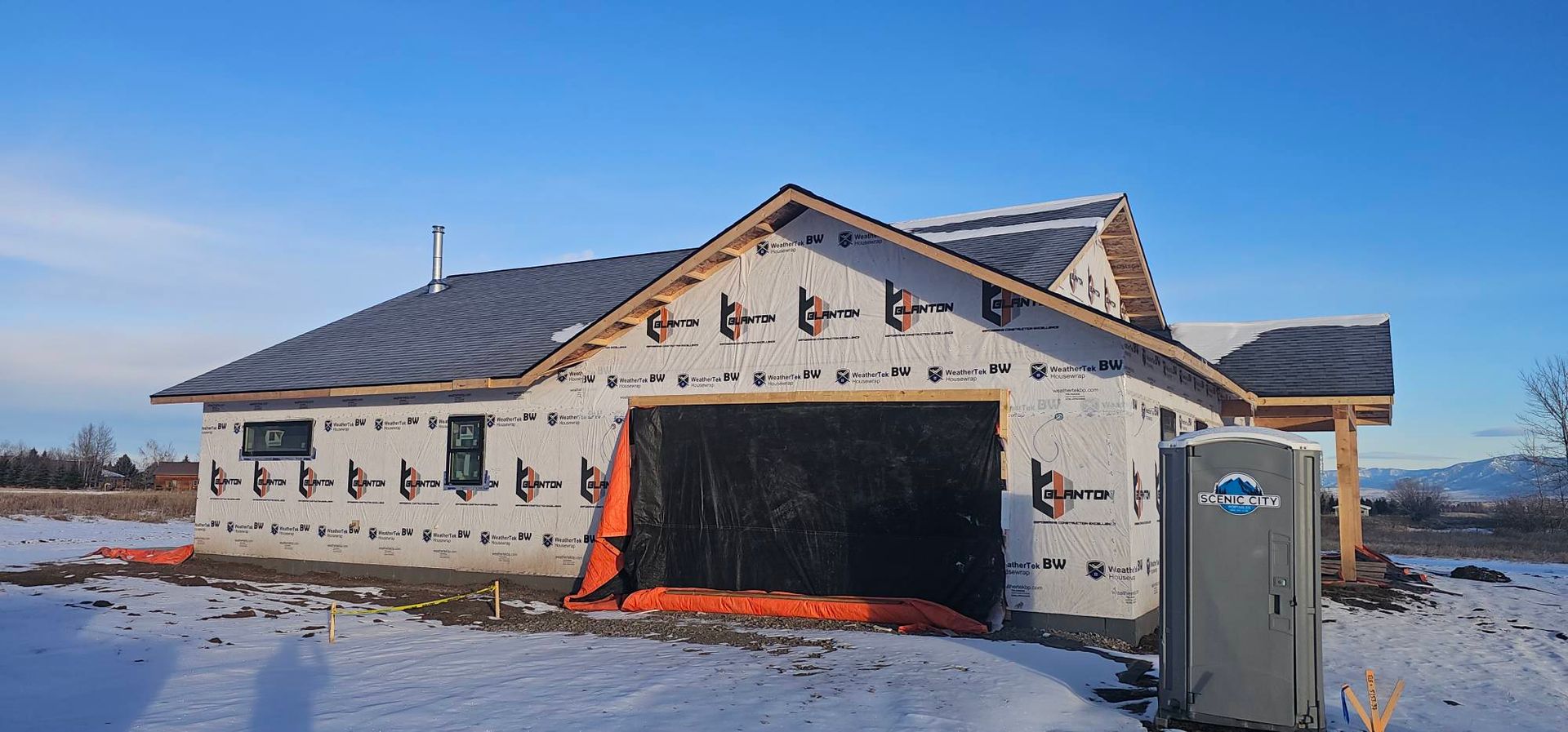 A house under construction with white exterior sheathing, a gray shingled roof, and a portable toilet on a snowy lot.