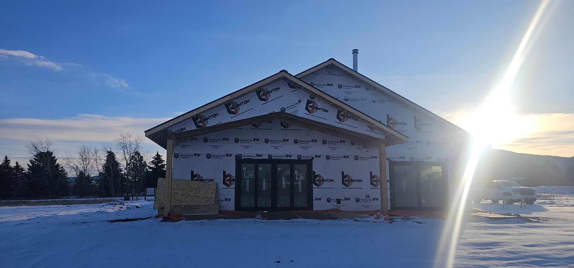 A house under construction with white exterior wrap, installed sliding doors, and a chimney, set in a snowy landscape.