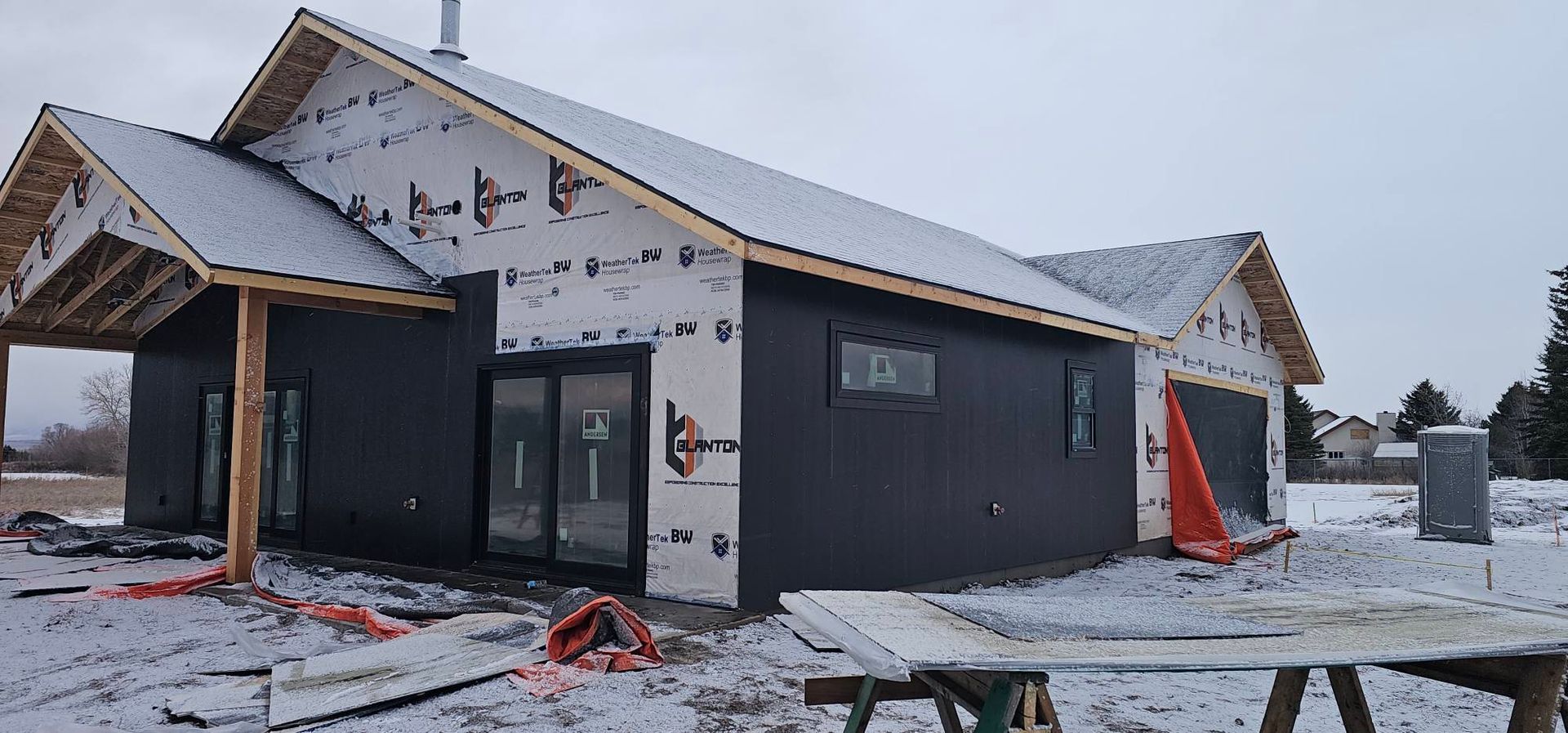 A house under construction with black siding, sliding glass doors, and a snow-covered yard on a cloudy day.