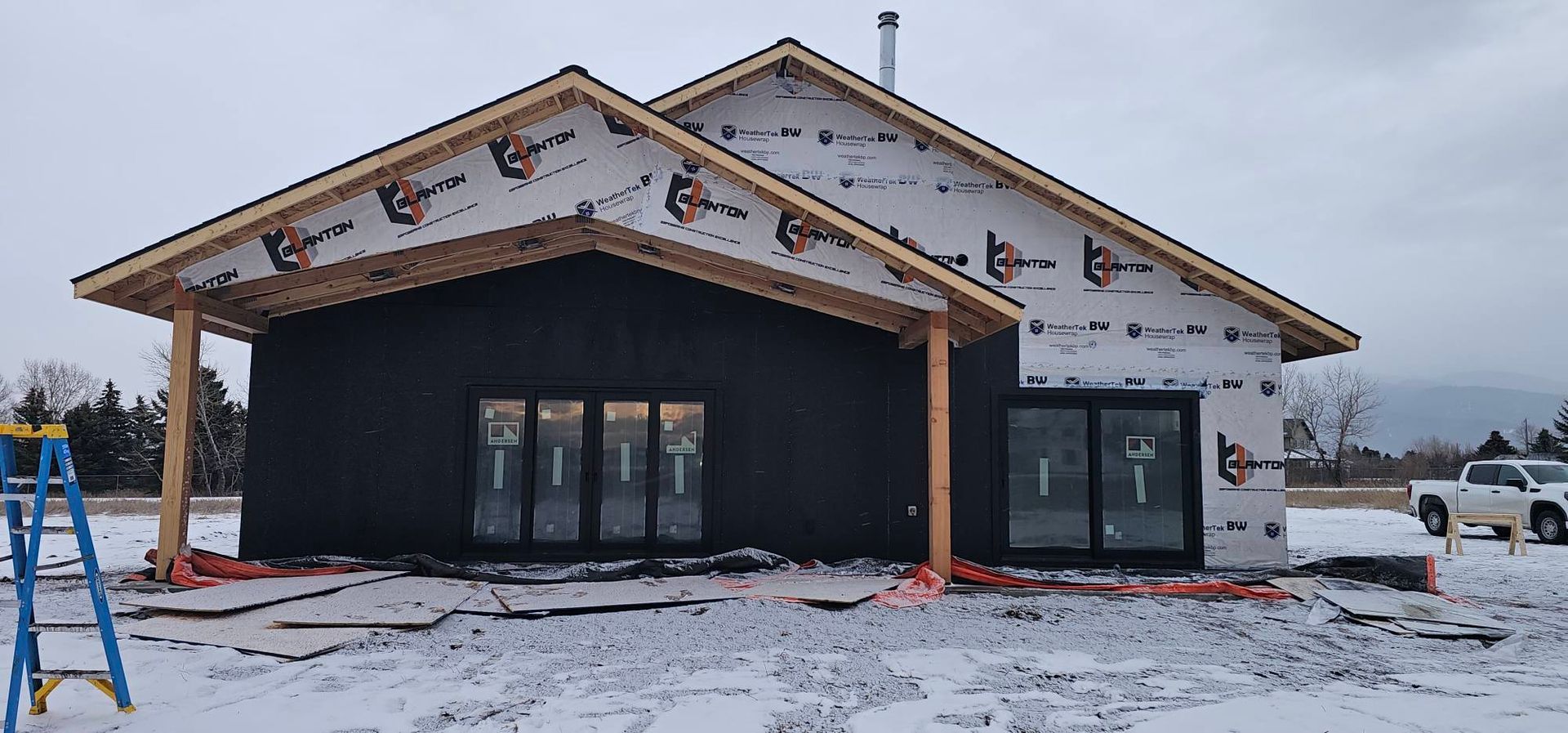 A house under construction with white building wrap on top, black siding below, large glass doors, and snowy surroundings.