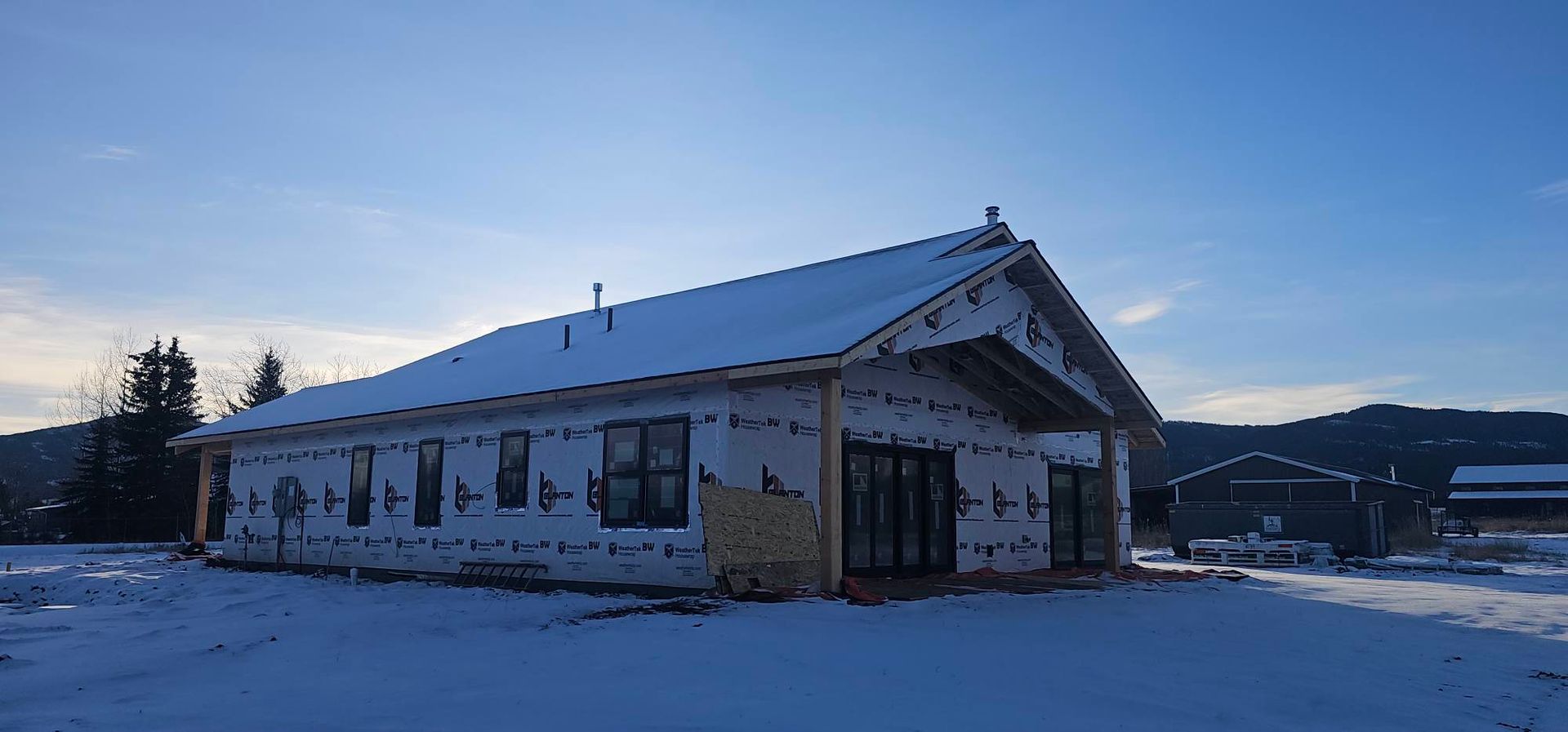 A house under construction with white exterior sheathing, black window frames, and a snow-covered roof at twilight.