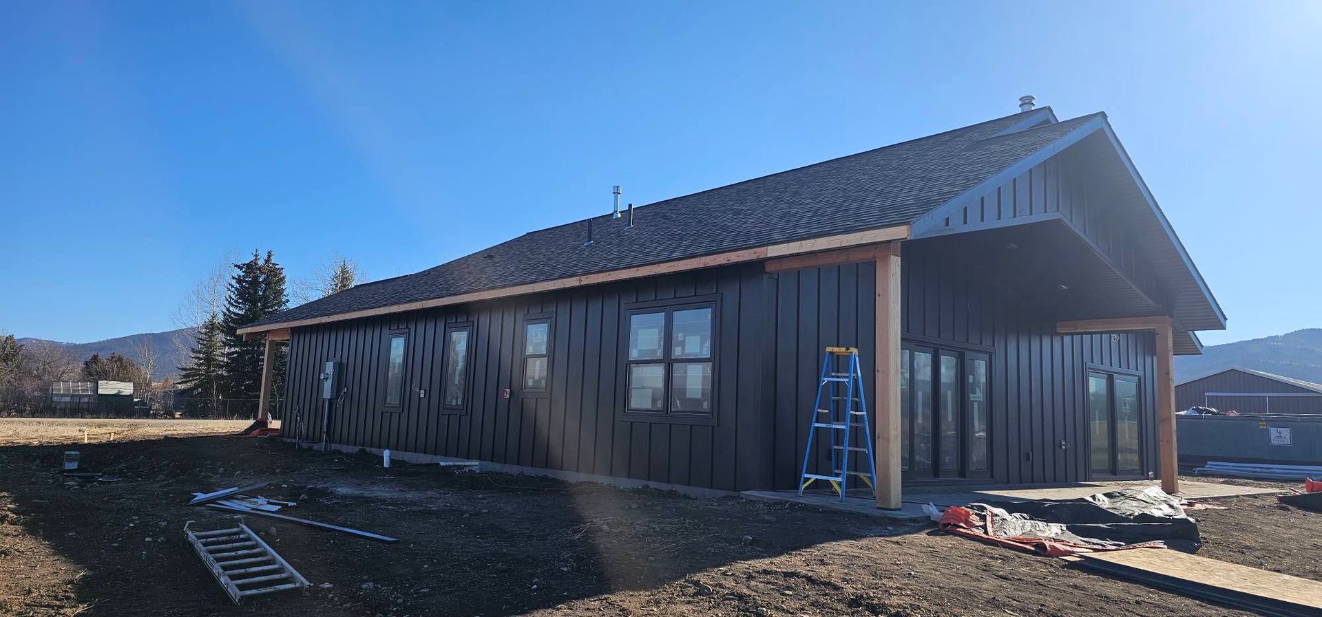 A modern single-story house with dark vertical siding and a gabled roof under construction on a sunny day.