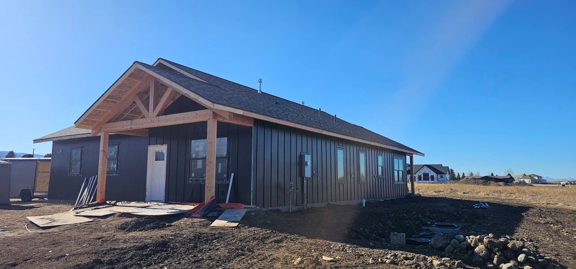 A single-story house under construction with black siding, a timber-framed front porch, and a clear blue sky.