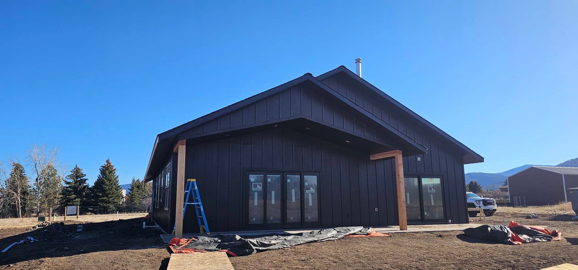 A newly constructed, dark-sided house with a covered patio, wooden support posts, and a ladder, set in a rural landscape.