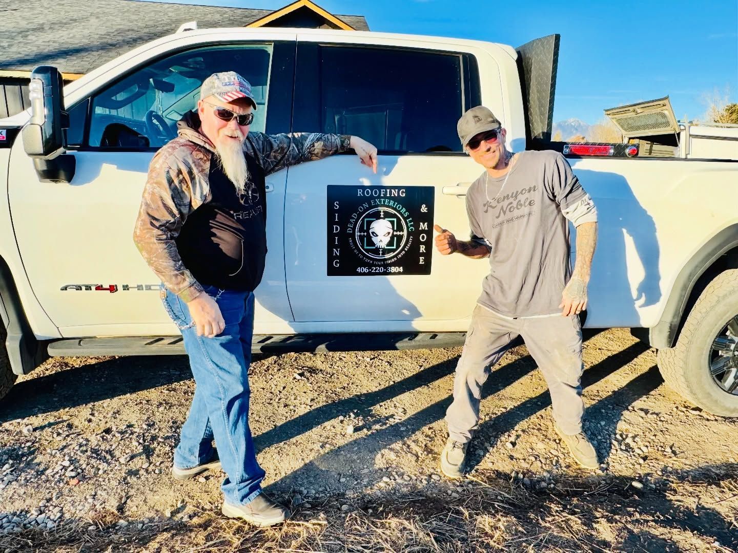 Two people stand next to a white truck in a dirt field, pointing at a square sign with an alien graphic on the door.