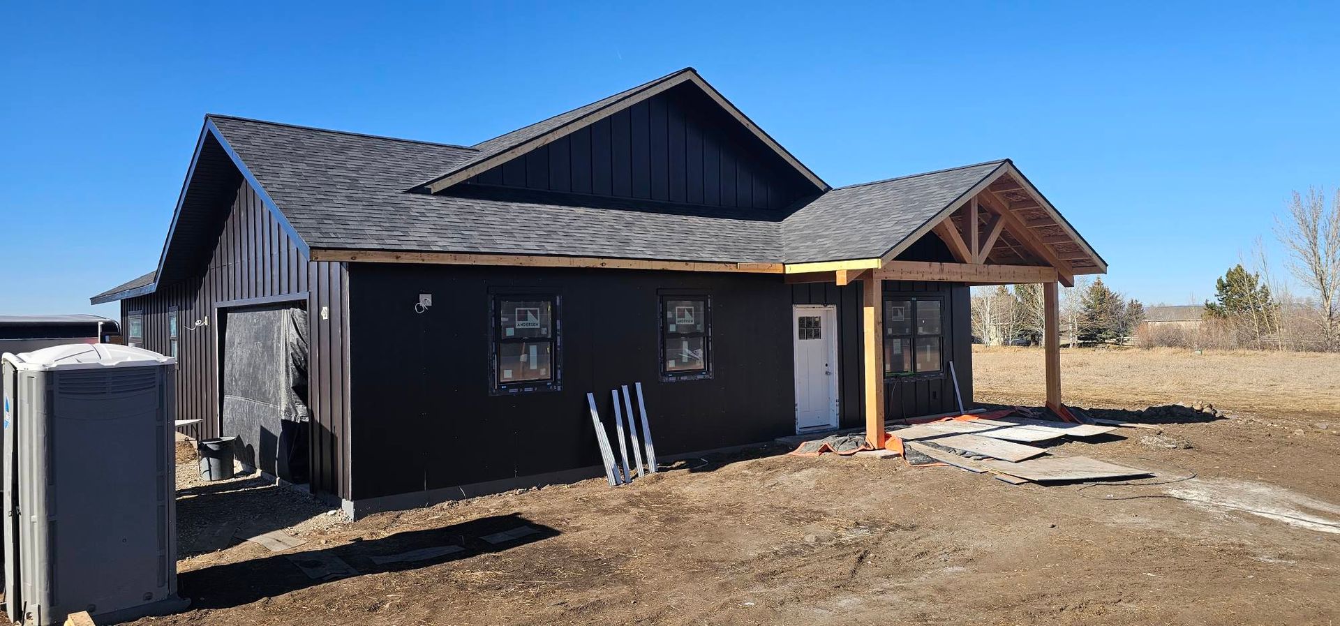 A dark-sided residential house under construction, featuring a prominent wooden porch frame and an adjacent portable toilet.