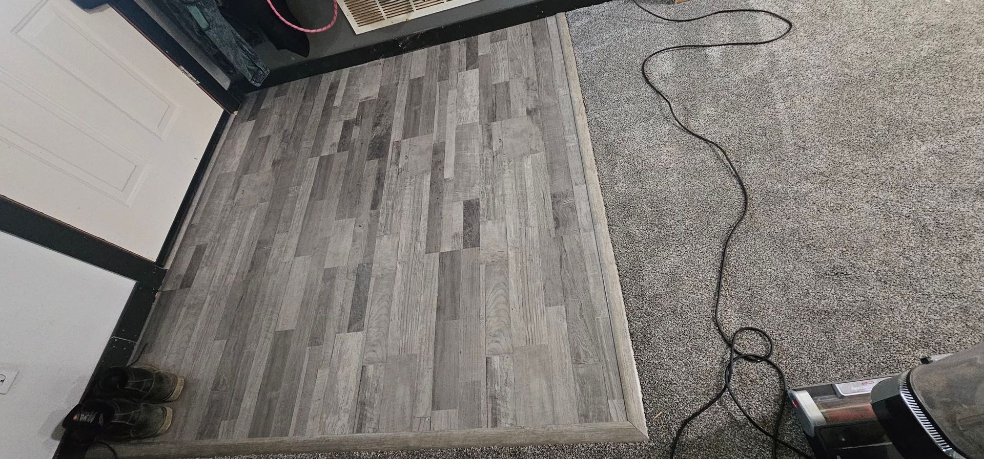 A view of a grey wood-plank floor transition into a grey speckled carpet in an indoor setting.