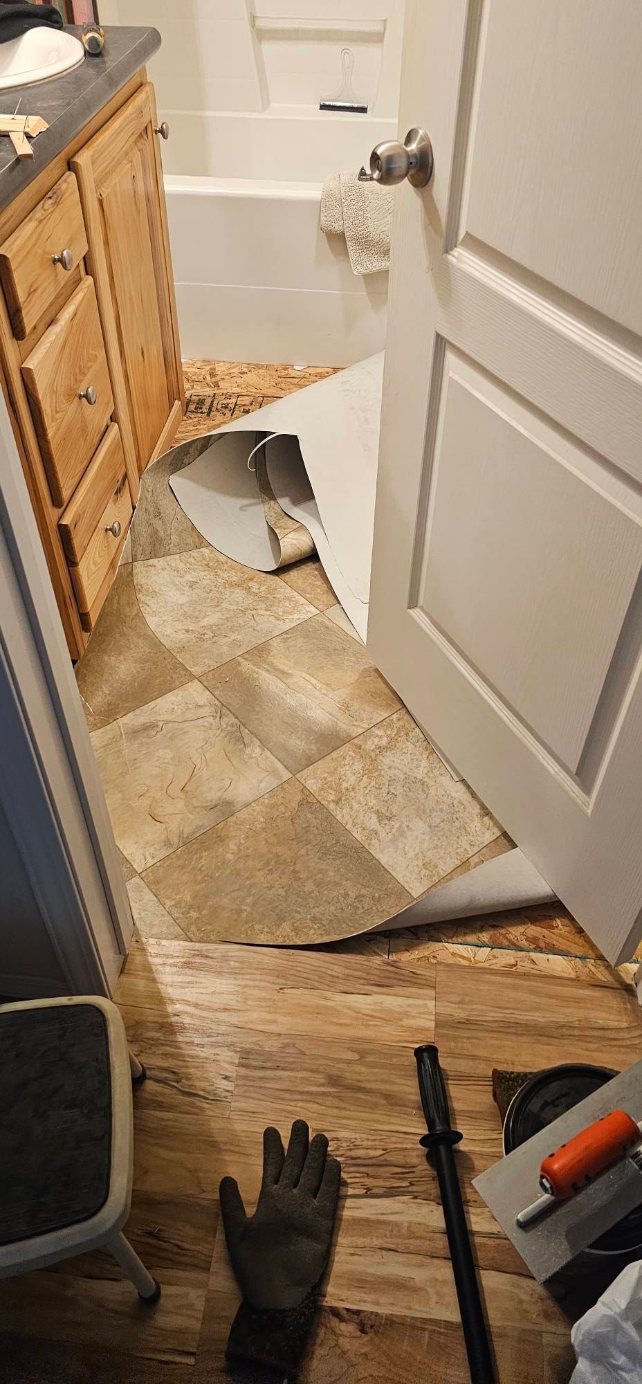 A bathroom under construction with peeling floor tiles and tools, including a glove and a trowel, on the floor.