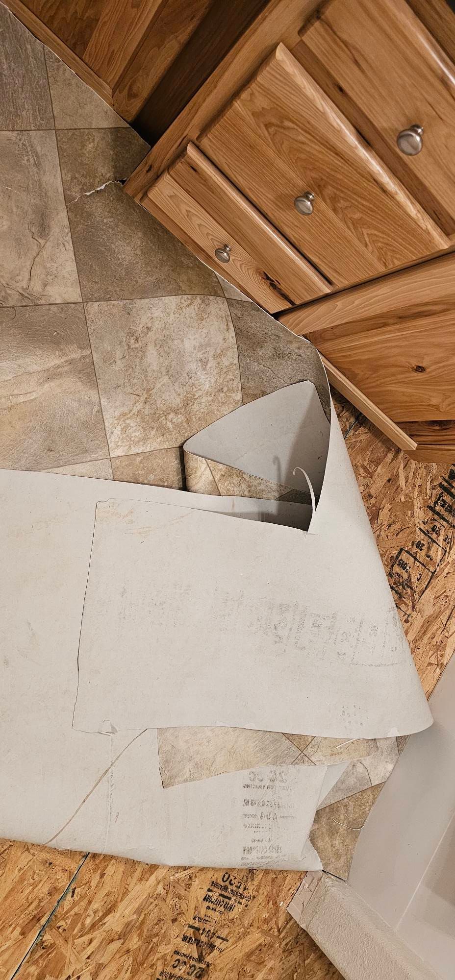 Damaged, peeling sheet vinyl flooring in a kitchen, revealing the wooden subfloor beneath near wooden cabinetry.