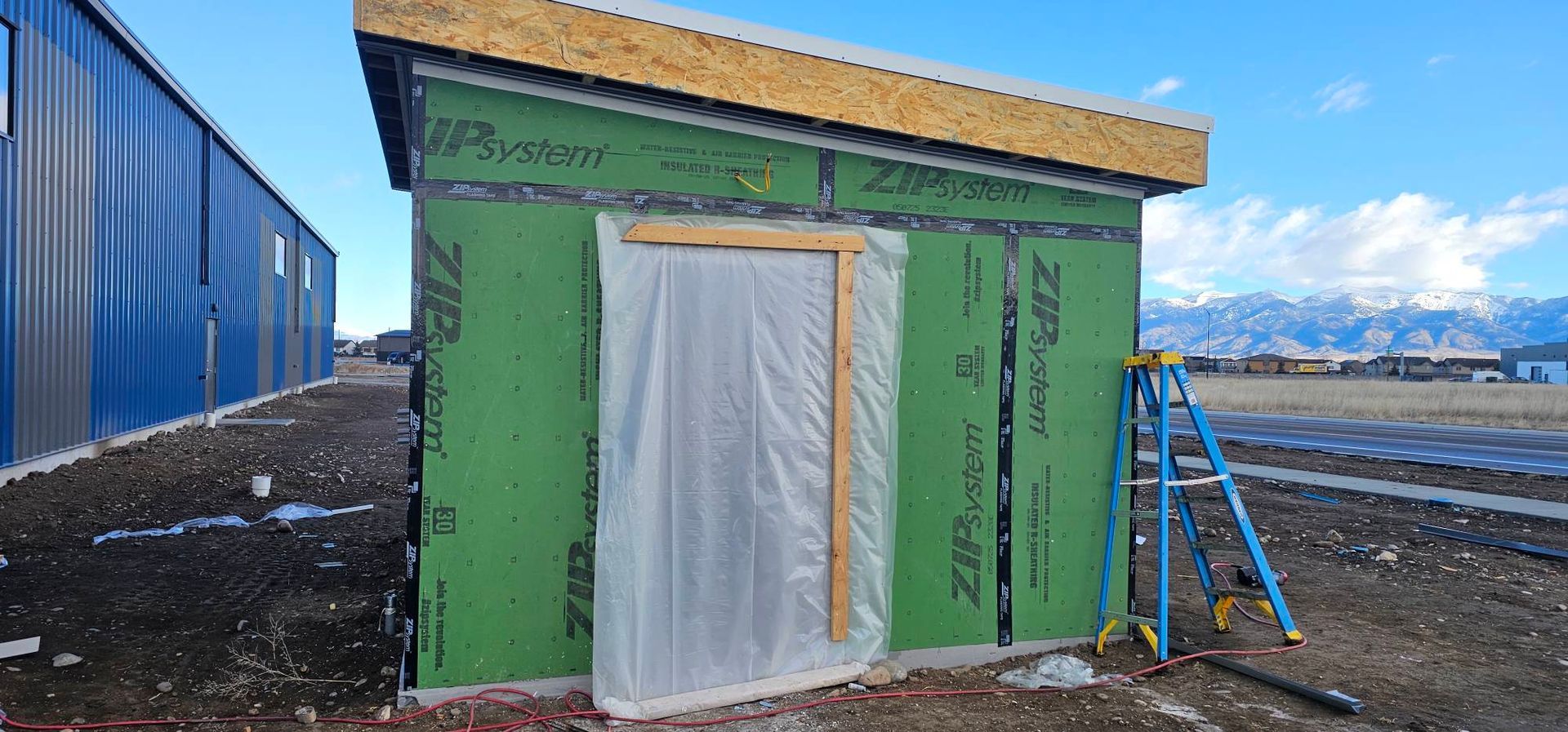 An unfinished small structure with green Zip System wall panels, a plastic-covered doorway, and a ladder on a dirt lot.