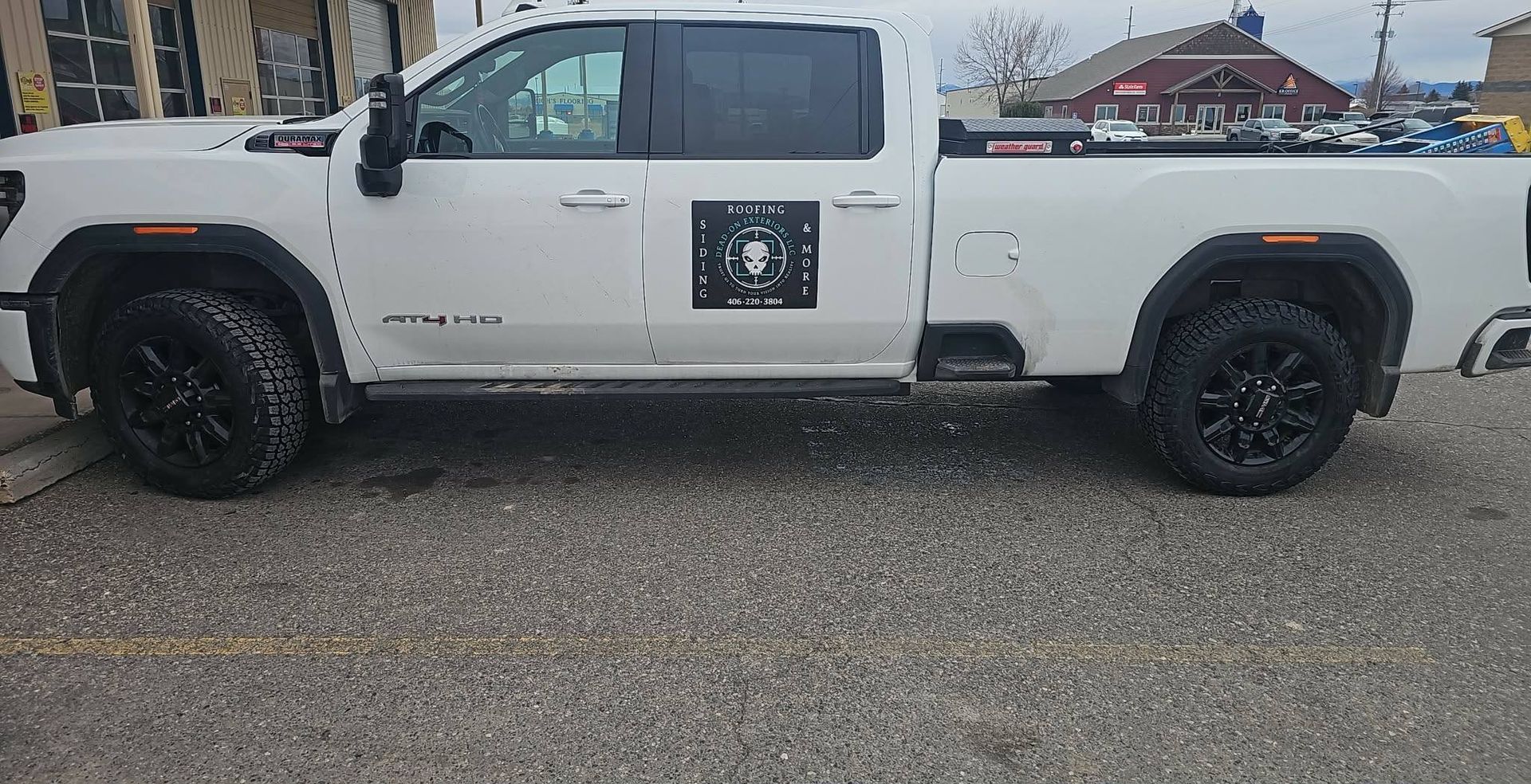 A white crew cab pickup truck with black wheels and a circular logo on the driver's side door, parked on a gravel lot.