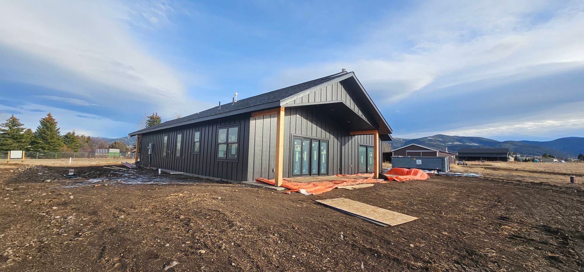 A newly constructed modern house with black vertical siding and a gabled roof, situated on a bare, dirt construction site.