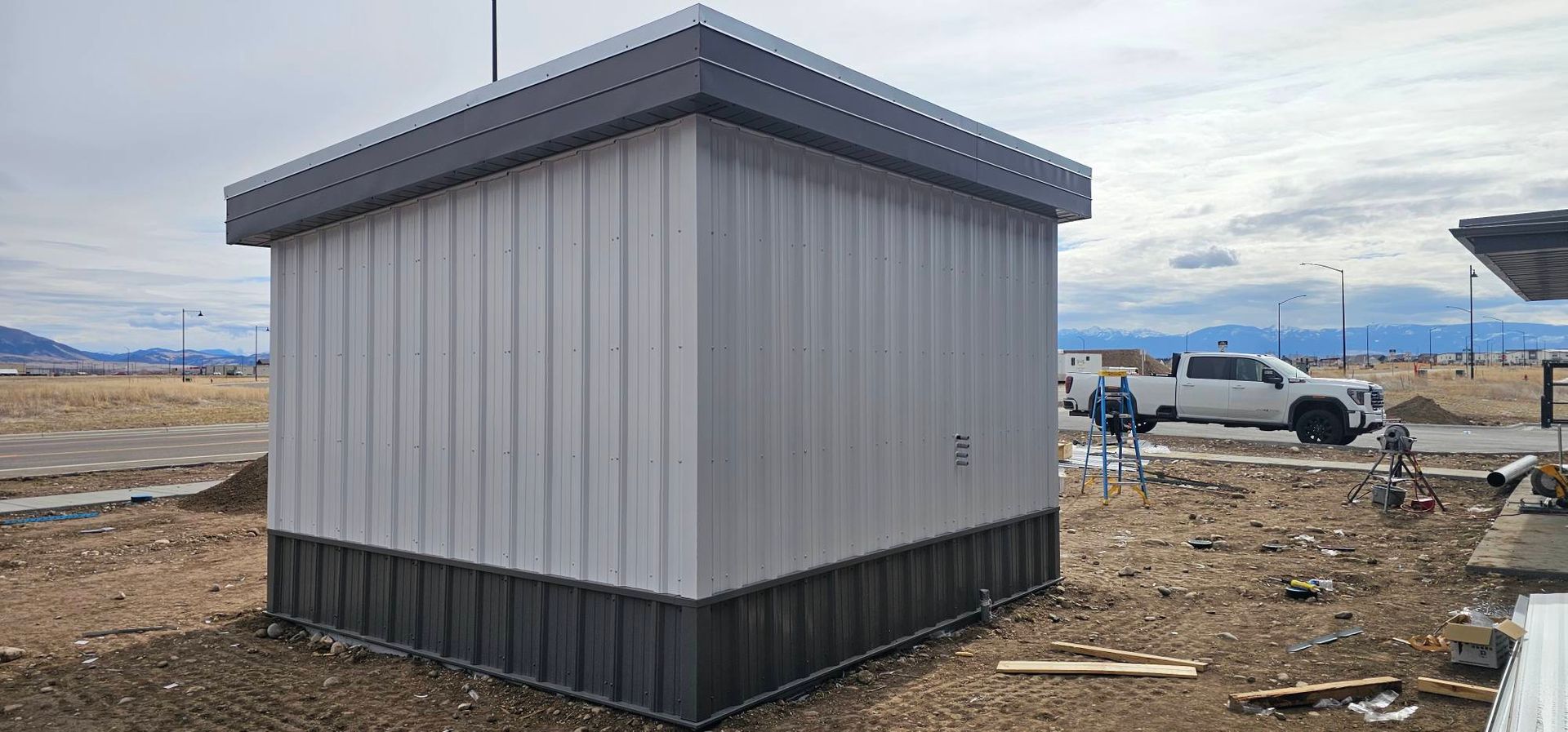 A rectangular metal utility structure with light grey walls, dark grey trim, and a matching base, set on a barren lot.