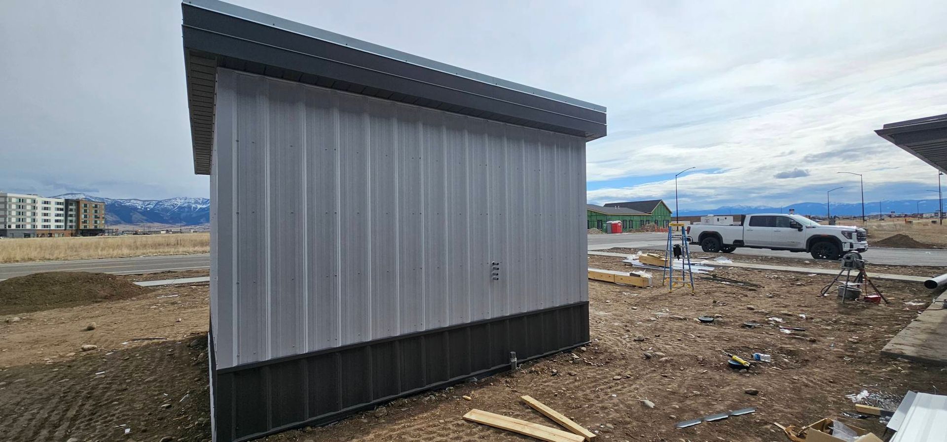 A small utility shed with white vertical siding and dark grey trim on a dirt construction site with distant mountains.