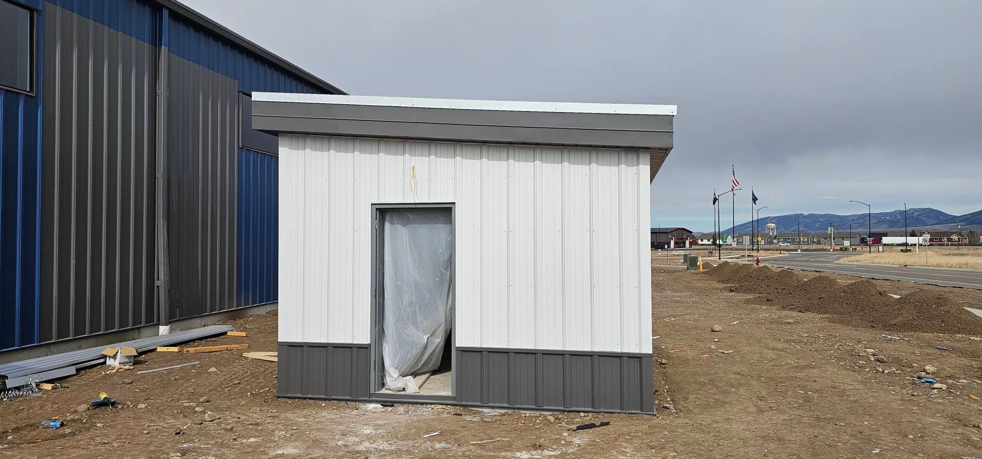 A small, white, rectangular metal shed with a gray trim and door, standing on a dirt lot beside a large warehouse.