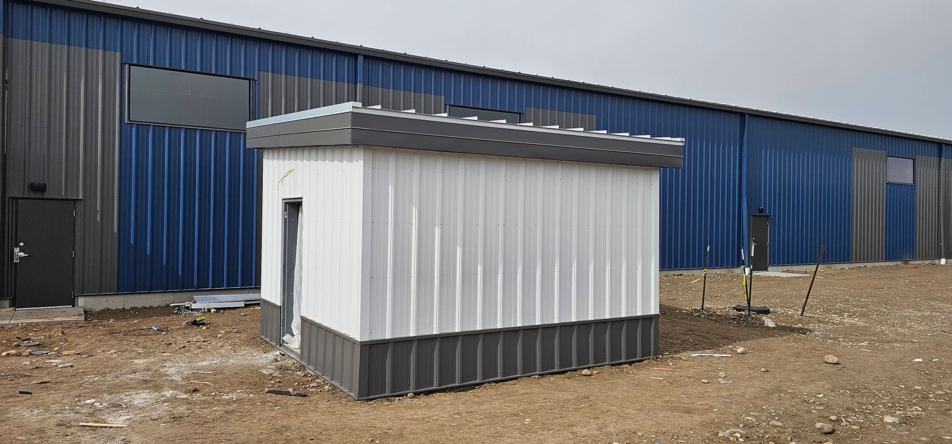 A small, white, rectangular metal building with a dark grey base and roof, standing in a field next to a large blue shed.