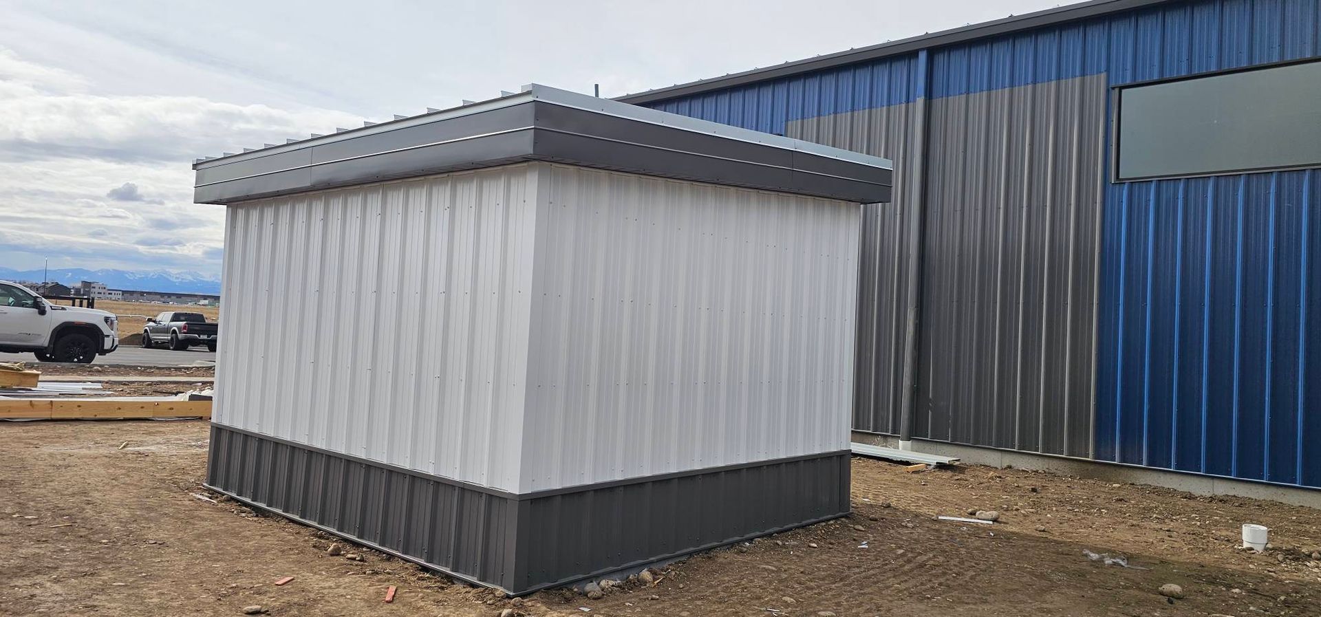 A small, square shed with white siding and gray trim, positioned next to a large blue and gray corrugated metal building.