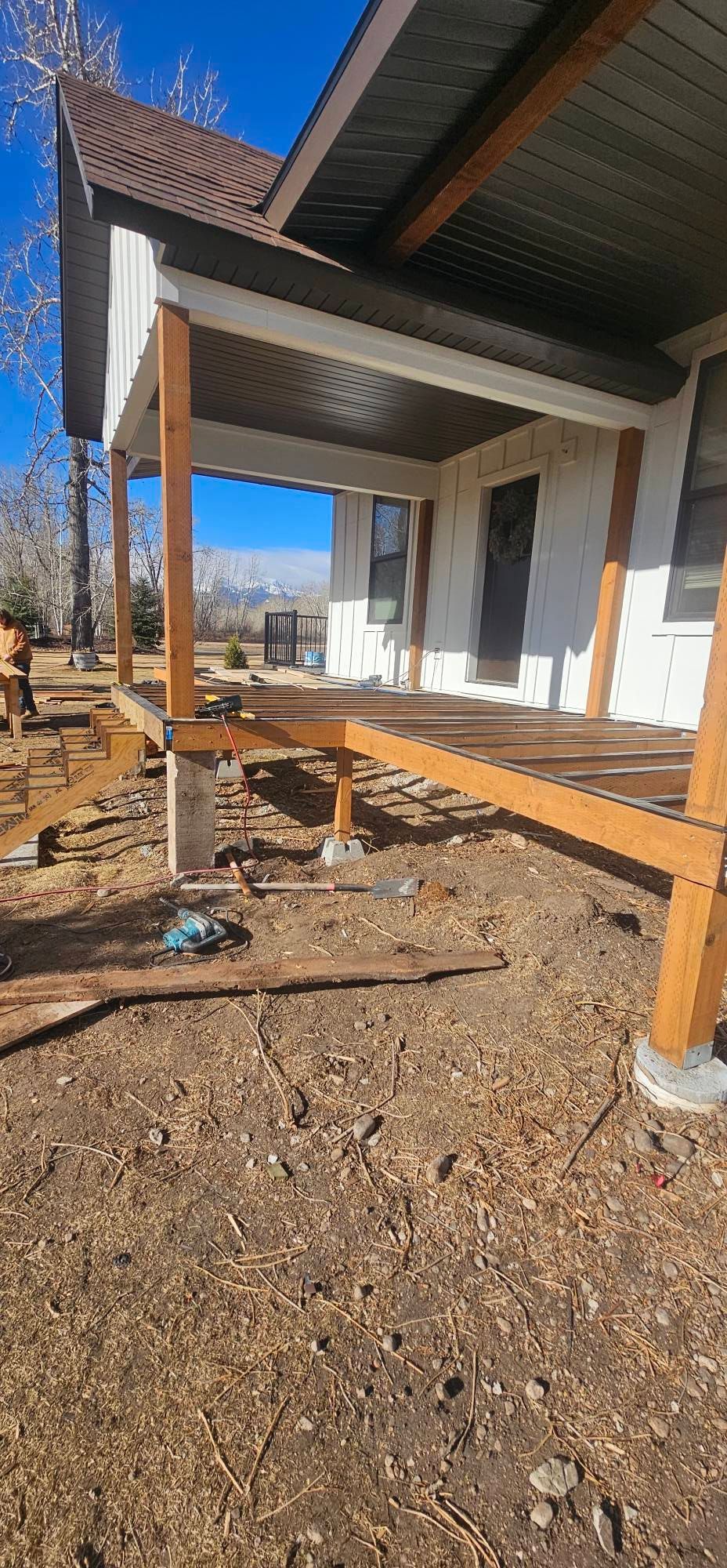 A wooden porch under construction attached to a house with white siding and a brown roof, set in a sunny, rural landscape.