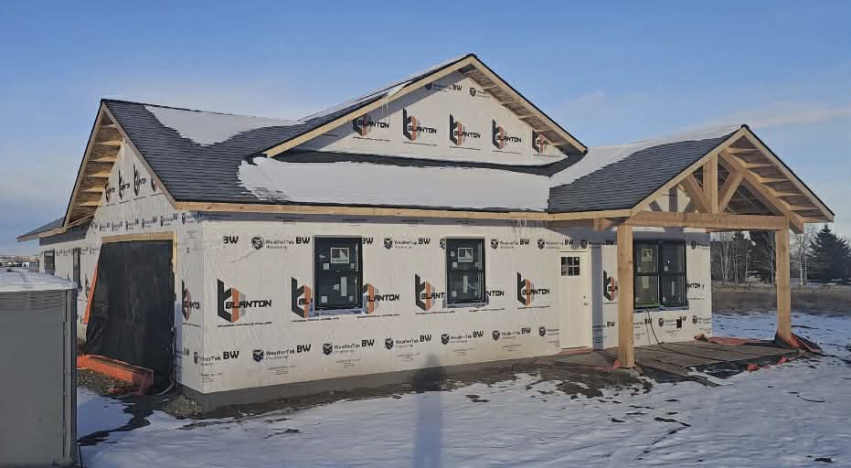 A newly framed house under construction with white exterior sheathing, black window frames, and a wooden porch structure.