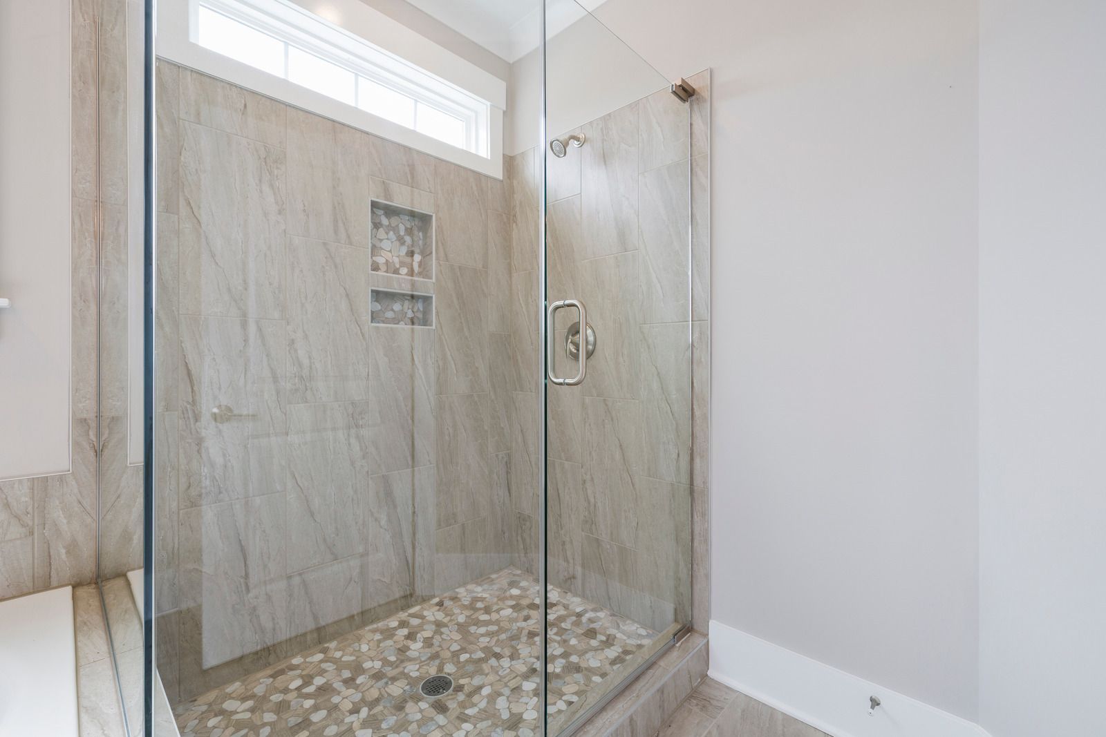A modern shower with glass doors, pebble floor, and light-colored tiled walls.