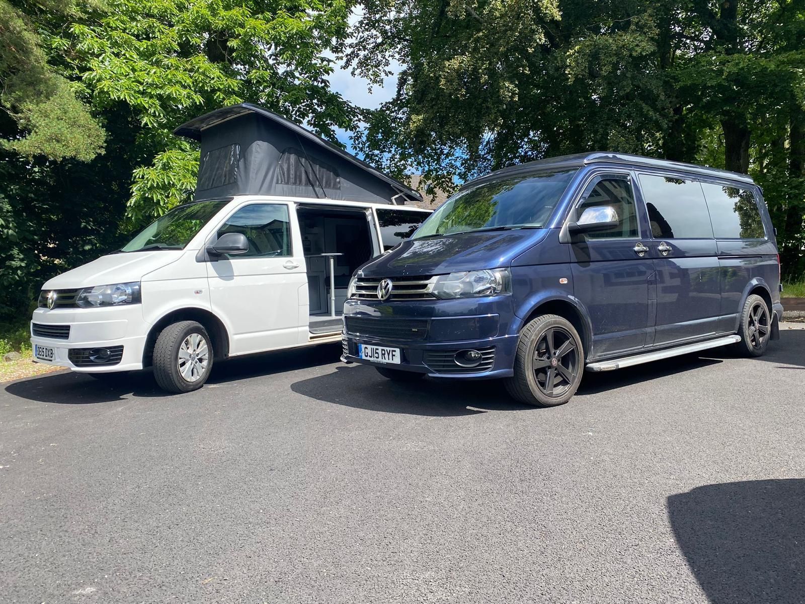 Two vans parked on a paved road, a white van with a westdubs pop top roof extended beside a dark blue van.