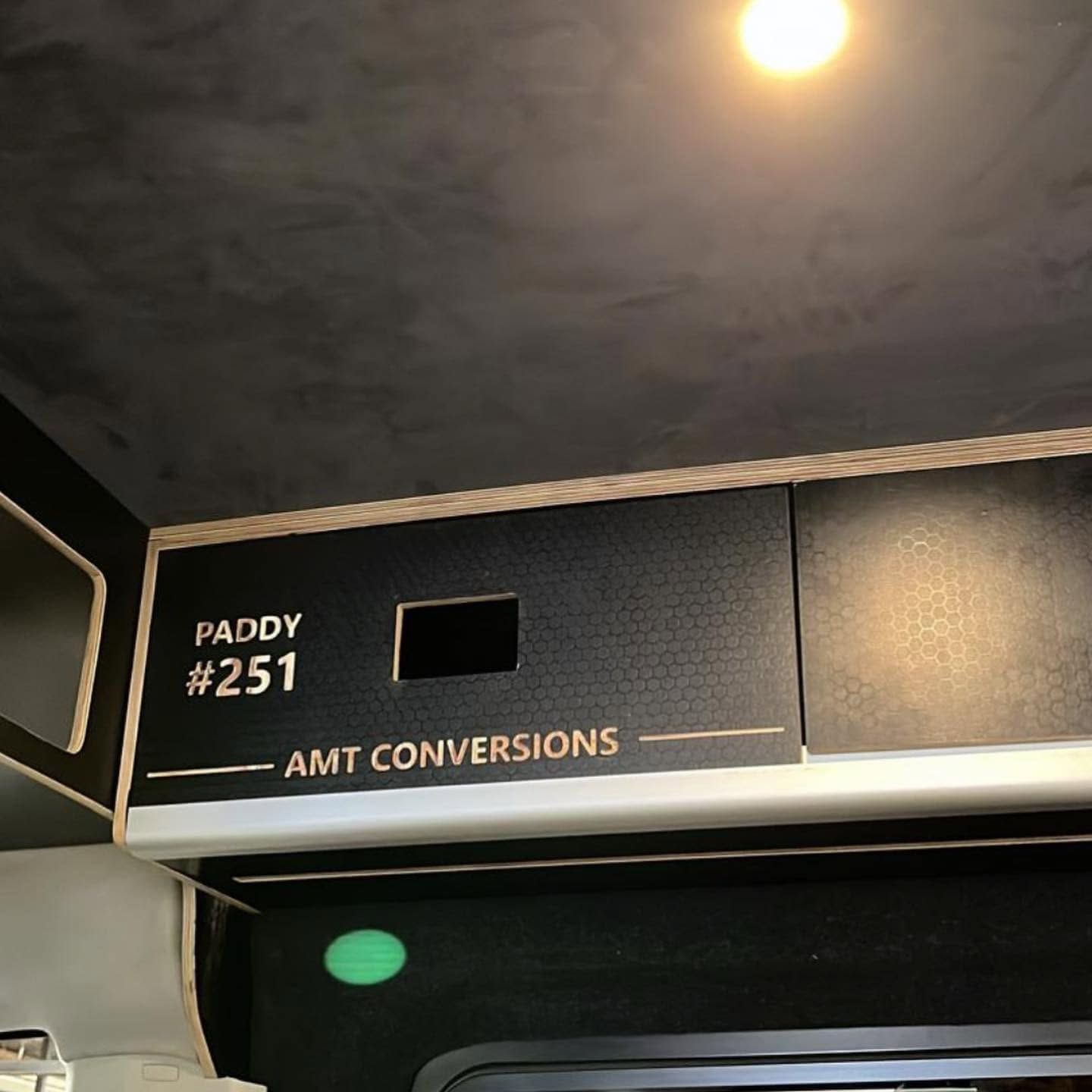 Airline overhead bin labeled “PADDY #251” and “AMT CONVERSIONS,” lit by a ceiling light.