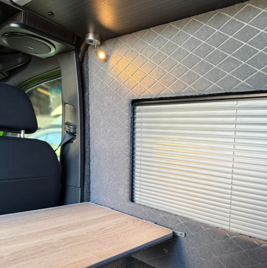 Van interior with quilted gray wall panel, window shade, and small wooden table beside a black seat
