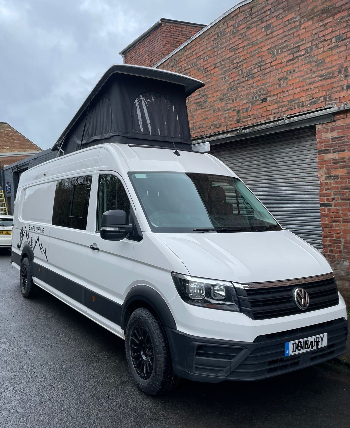 White Volkswagen van with raised pop-top roof parked beside a brick building