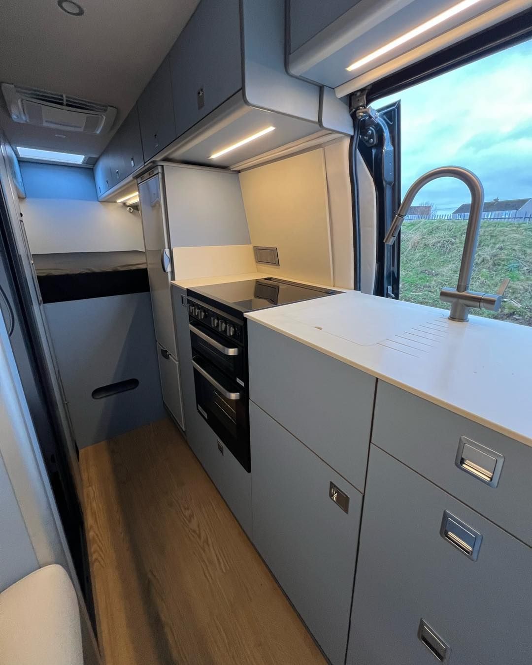 Compact camper van kitchen with gray cabinets, sink by window, and wood floor.