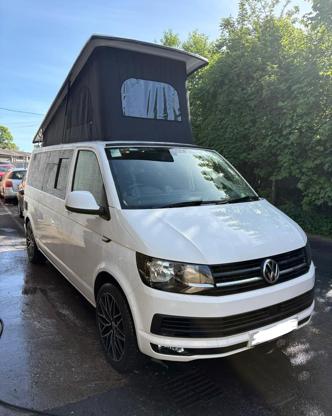 White Volkswagen van with pop-top roof parked on a wet street beside trees