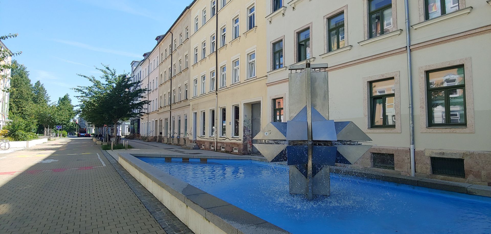 Sunlit quiet street with a long beige building and a large blue star sculpture beside a shallow reflecting pool
