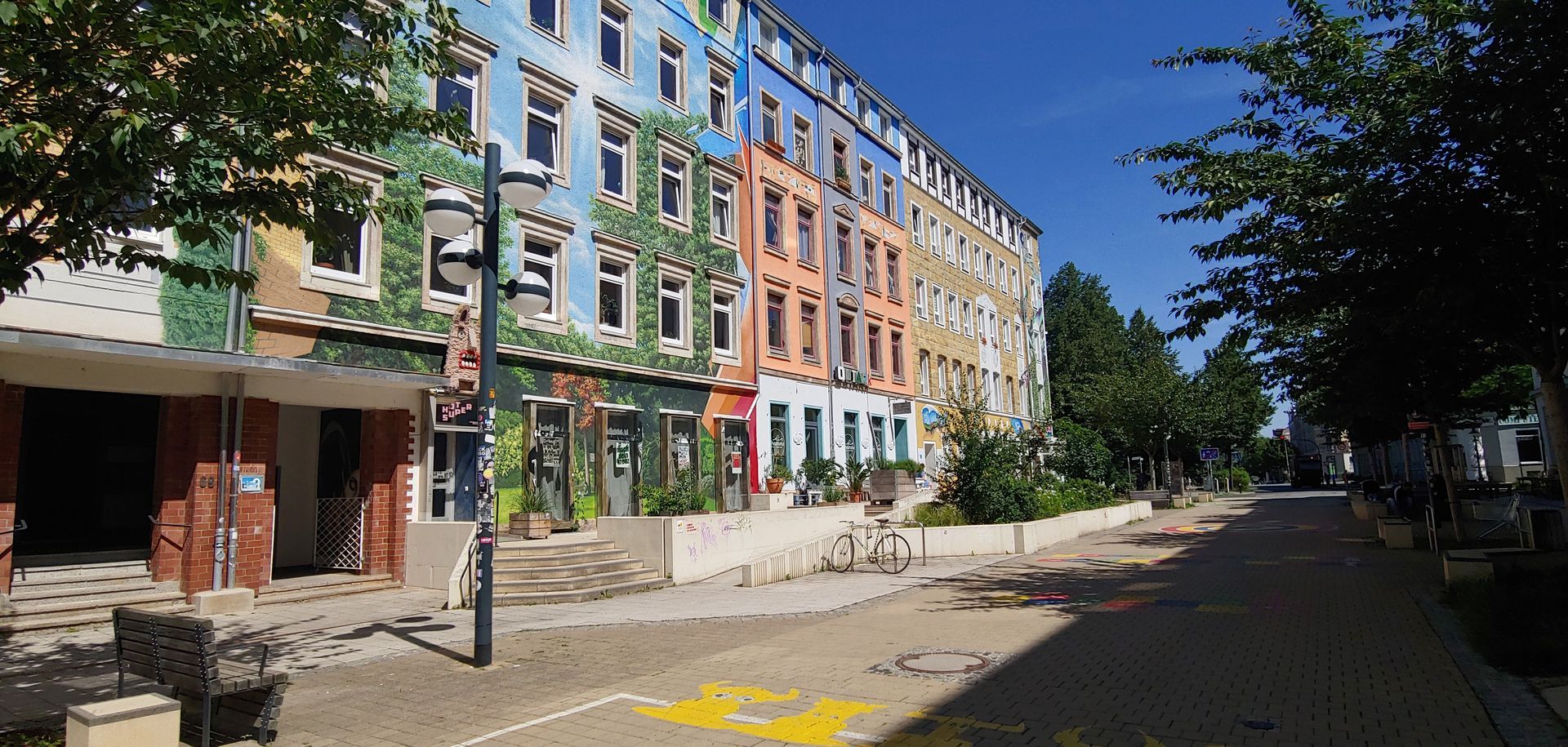 Tree-lined street with colorful apartment buildings, brick sidewalk, and parked cars under a clear blue sky