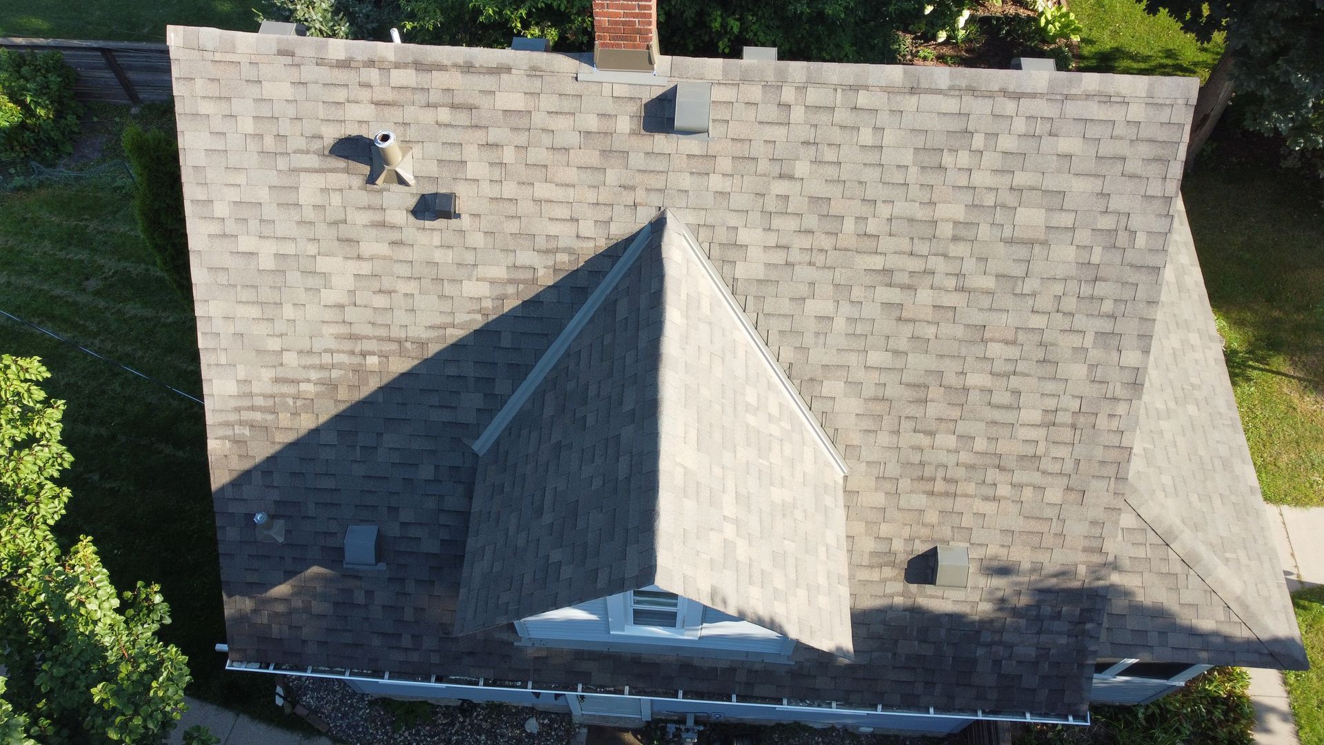Overhead view of a house roof with grey shingles, chimney, and dormer in a grassy setting.