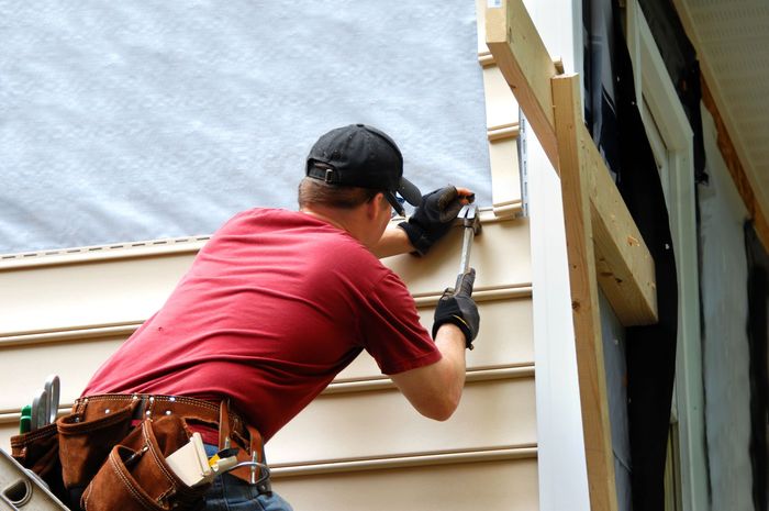 Person in a red shirt installing siding on a building's exterior, using a hammer and wearing a tool belt. Person in a red shirt installing siding on a building's exterior, using a hammer and wearing a tool belt.