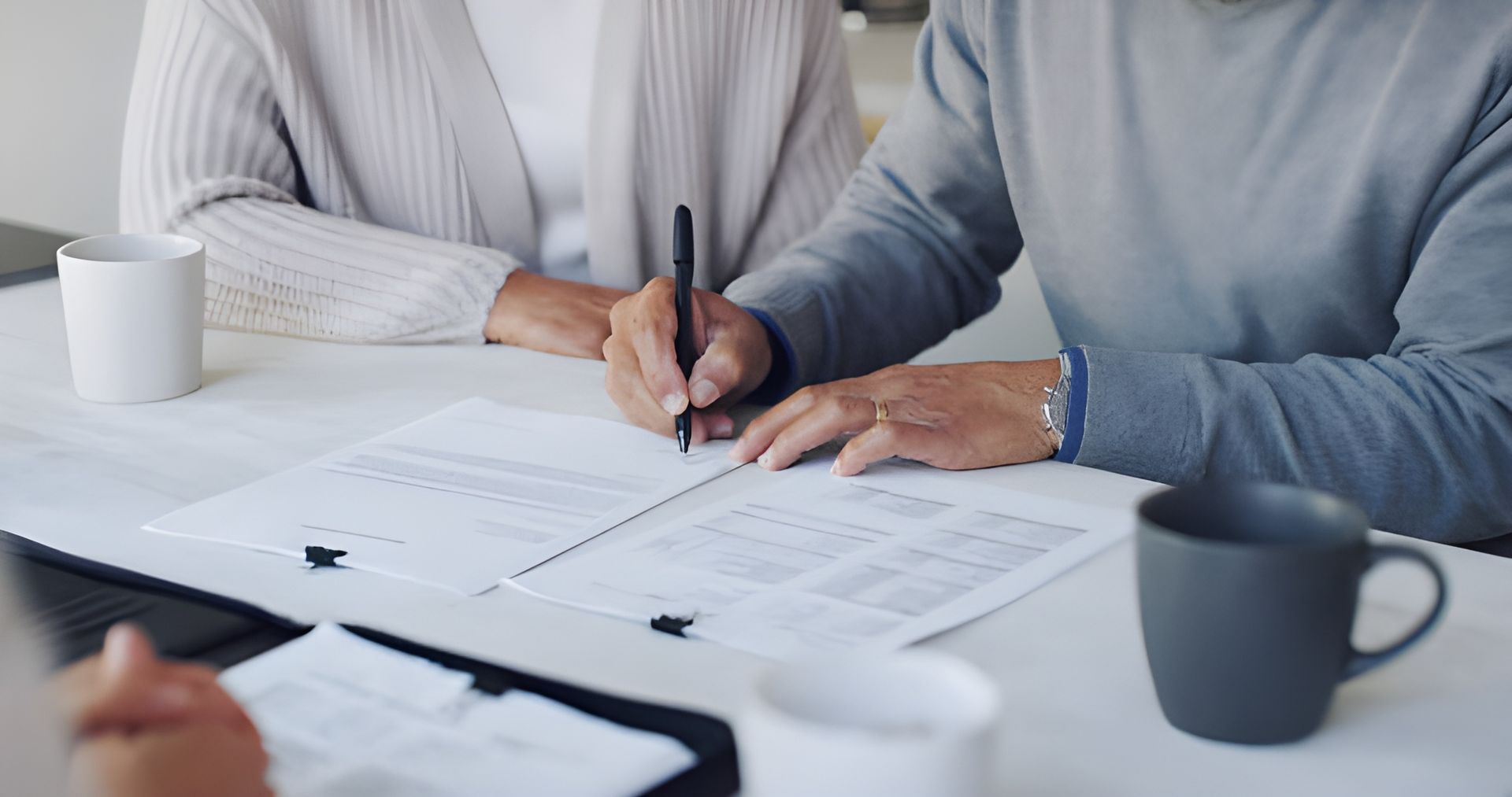 Person signing a document, seated at a table with another person, mugs, and papers.