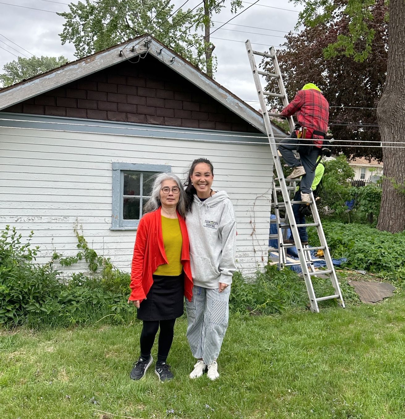 Two women pose in front of a white building with someone on a ladder working on electrical lines.