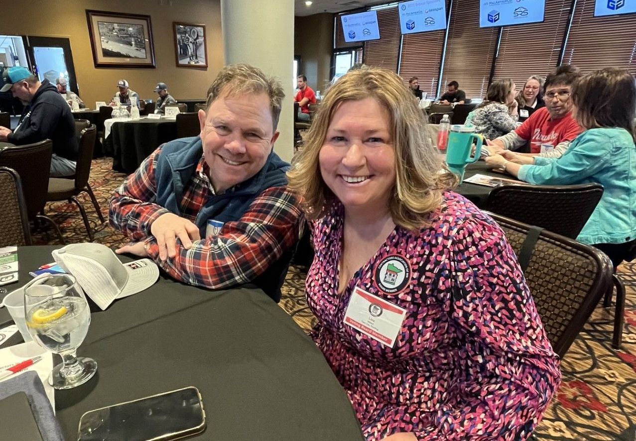 Two smiling people sit at a table in a conference room. The woman wears a pink patterned shirt, the man a plaid shirt.