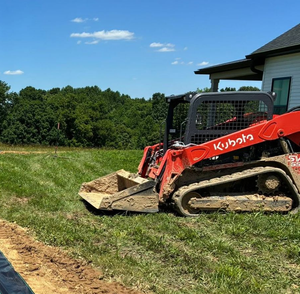 Red Kubota compact excavator grading a grassy yard beside a house under a blue sky