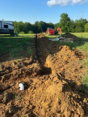 Large dirt trench in a grassy field with a backhoe and excavator working near a white RV.
