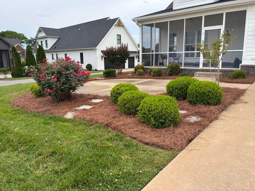 Landscaped front yard with rounded shrubs, pink flowers, and a white house porch in the background
