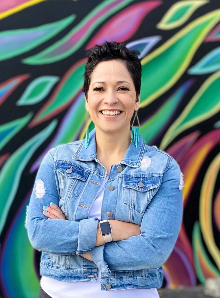 Woman with short hair, smiling, arms crossed, wearing denim jacket, standing in front of a colorful mural.