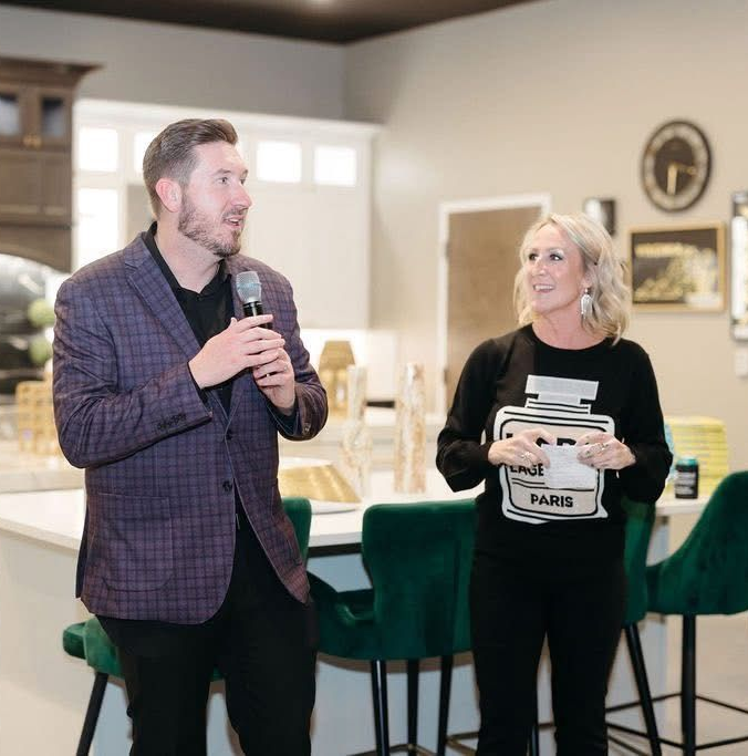 Two people speaking indoors; man holds a microphone, woman holds a PARIS sign and smiles.