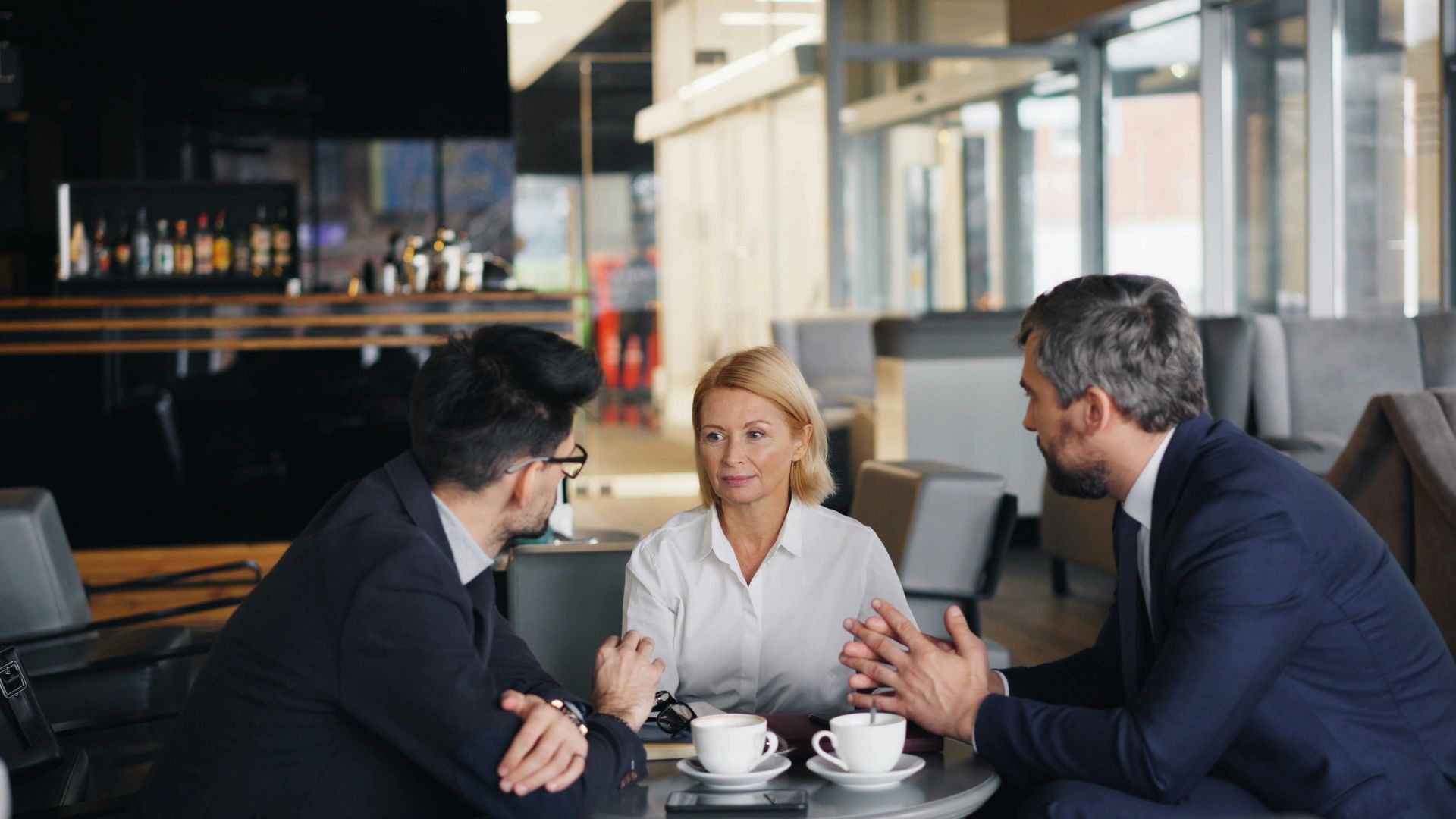 Three colleagues in business attire discussing over coffee at a table in a modern office lounge.